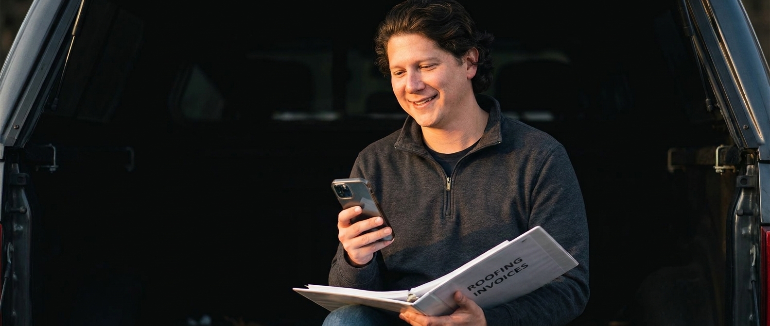 Vincent Jordan on a truck tailgate with a roofing invoice binder, checking his phone
